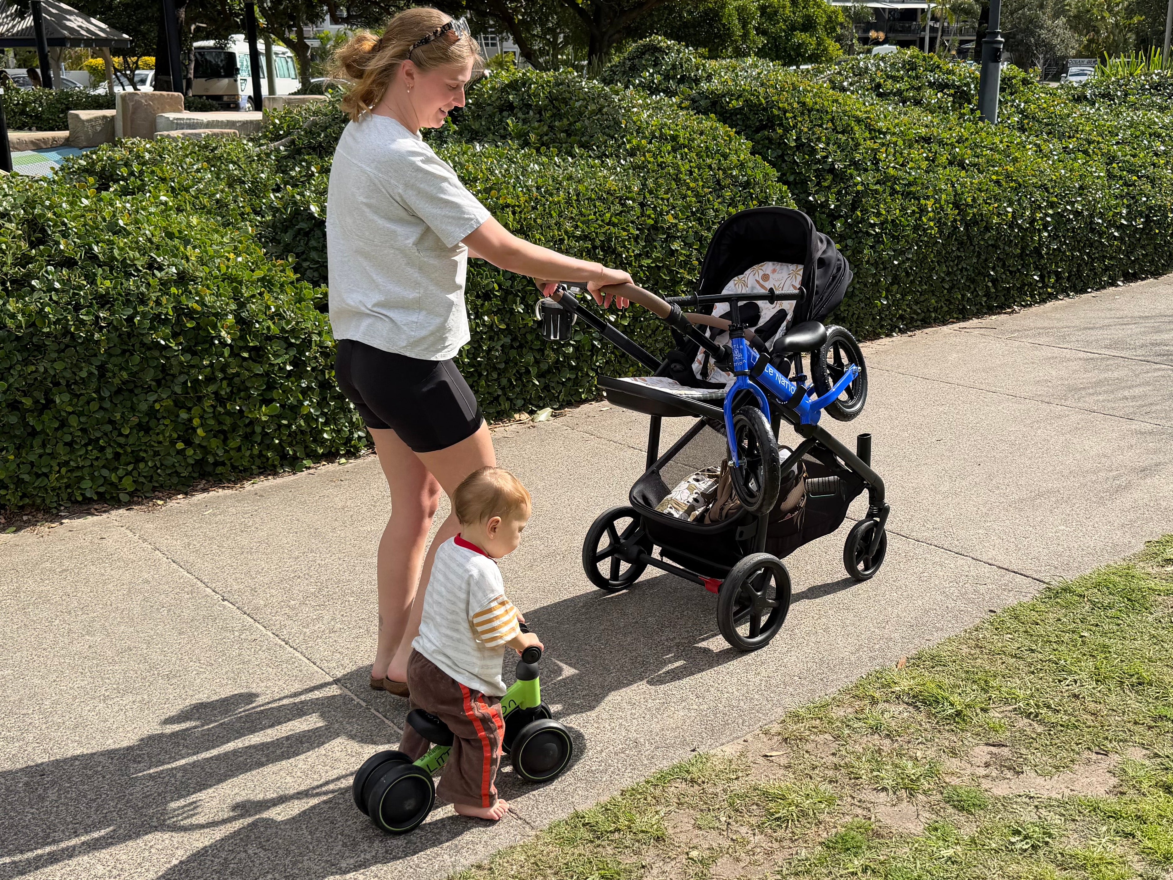 A woman pushes a black and blue stroller while walking on a paved path next to a child on a small green tricycle. The background features a green hedge and outdoor park setting.